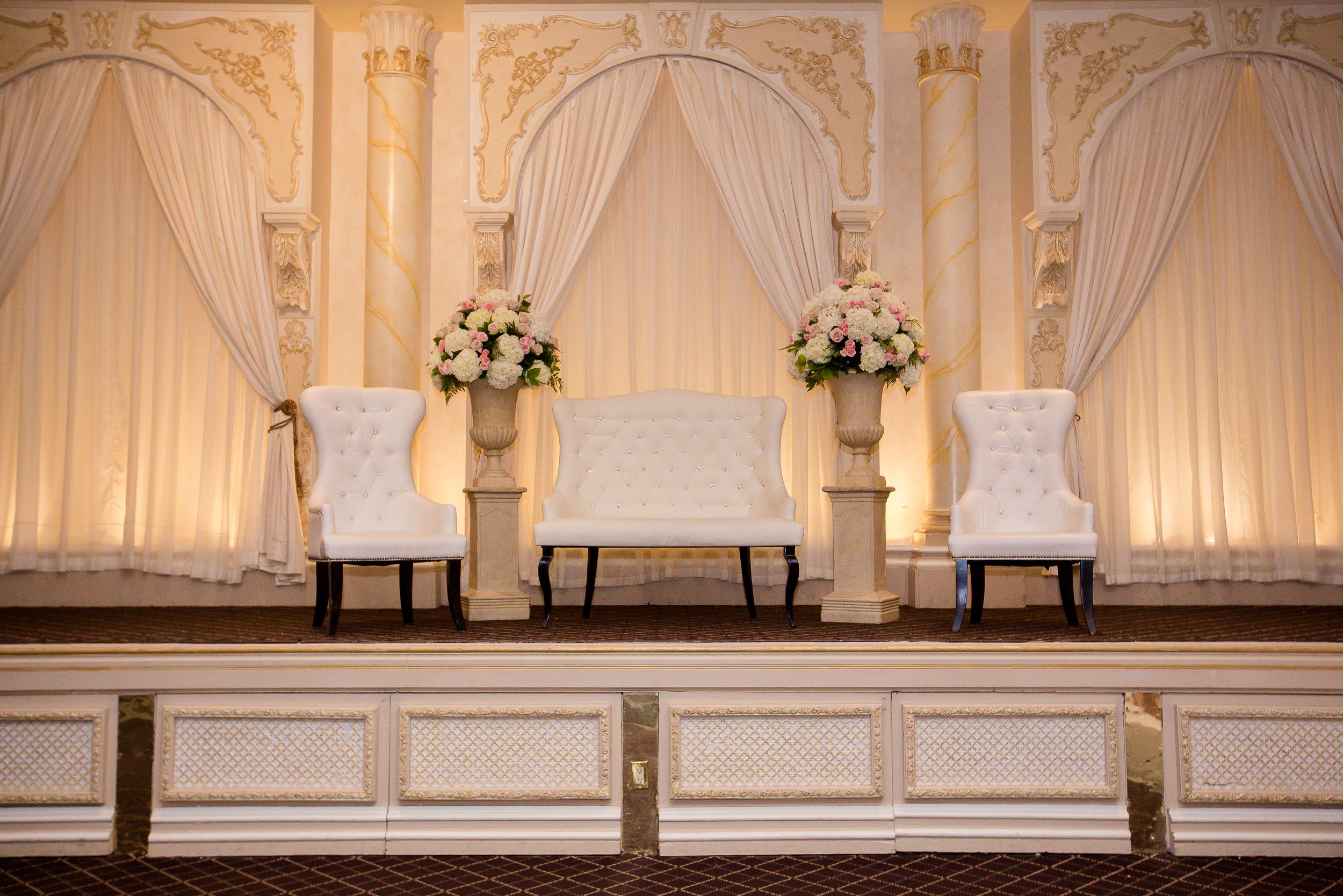 Luxurious wedding stage with white chairs, floral arrangements, and draped backdrop